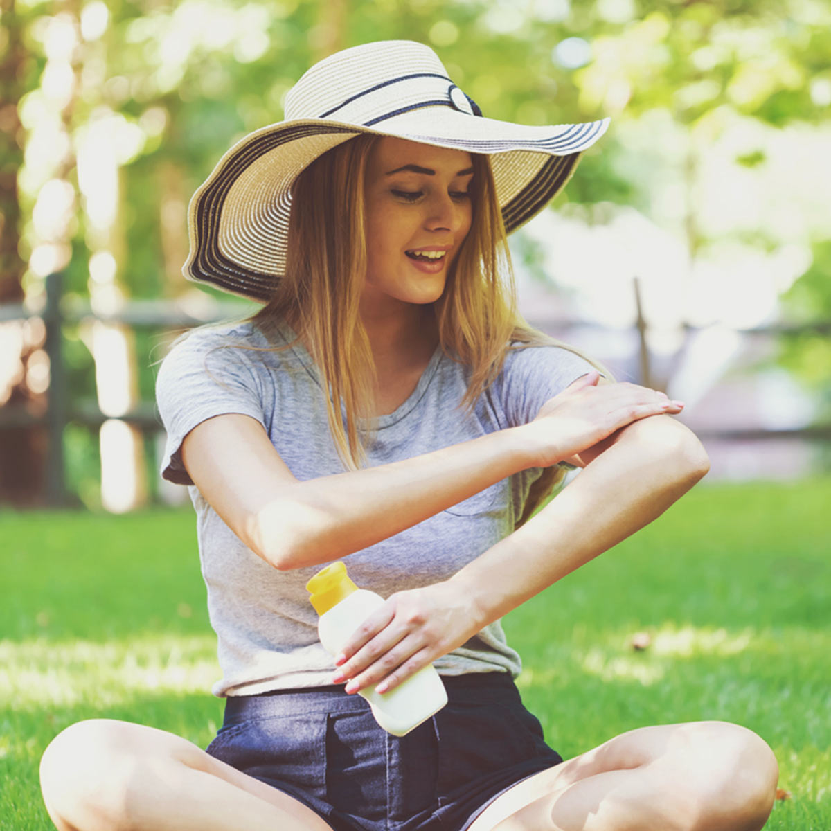 A young woman sits cross-legged in a green park on a sunny day. She is wearing a wide-brimmed hand and applying sunscreen lotion.