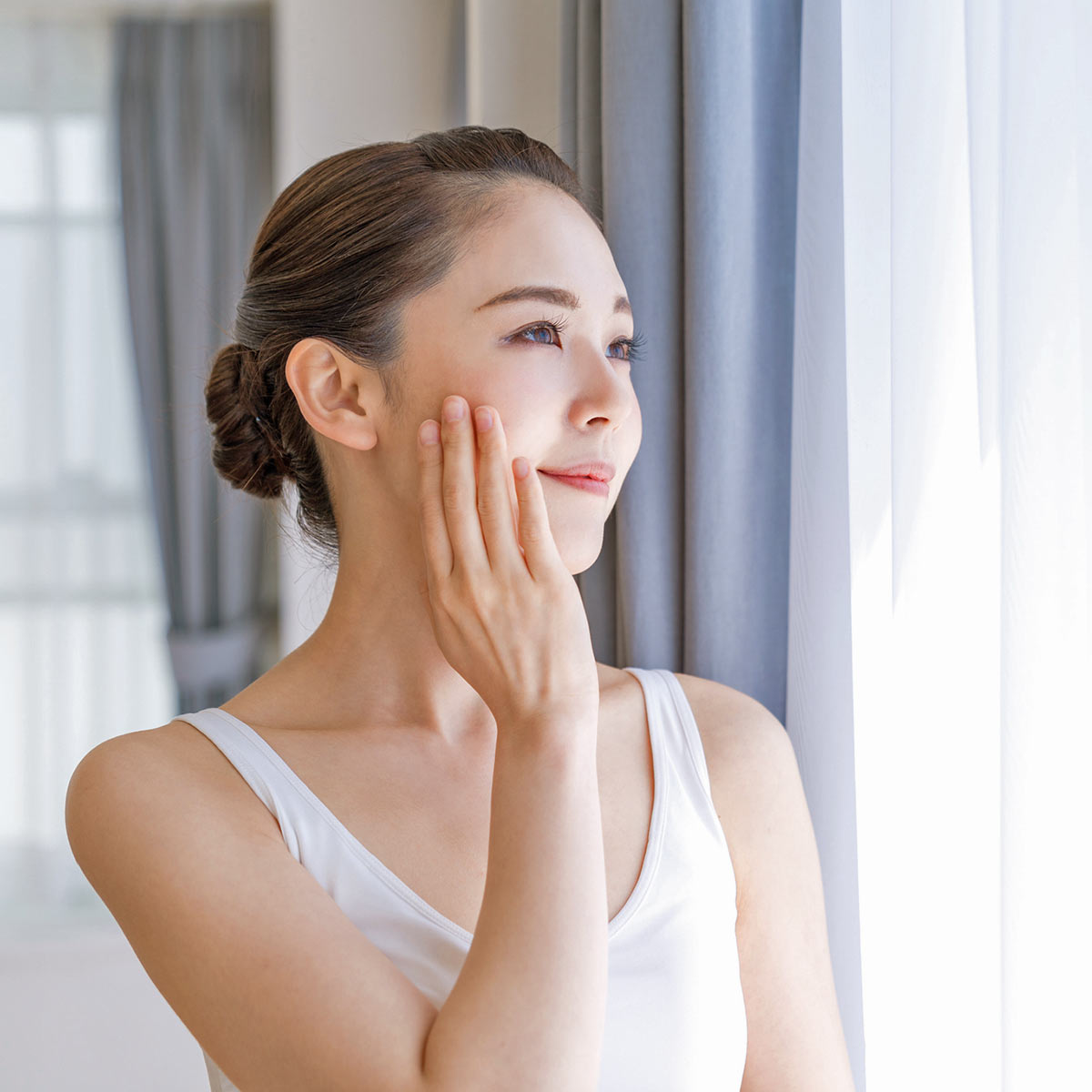 A young asian woman in a tank top looks out a window while smiling thoughtfully.