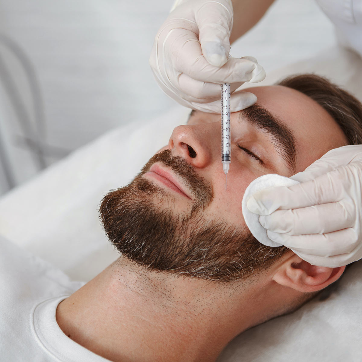 A bearded white man lies on a treatment table while he receives a dermal filler injection to his cheek.
