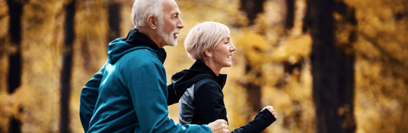 A man and a woman of retirement age jog together in a forest with fall foliage.